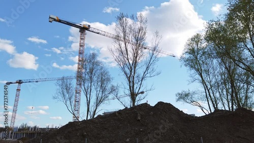 Cranes at a construction site against a blue sky, with a pile of dirt in the foreground