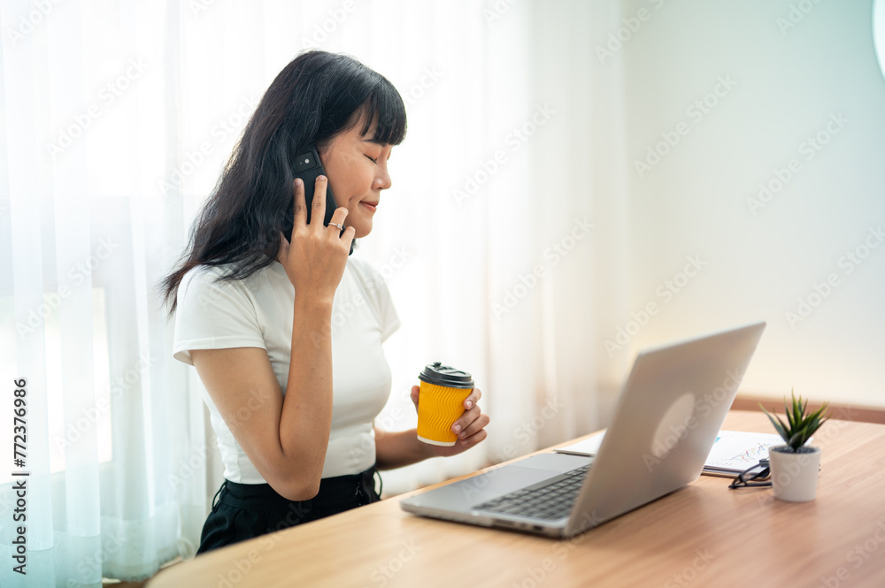 Asian woman talking on smartphone while enjoying with coffee and working on laptop In a bright room with white curtains Asian woman talking on smartphone while enjoying with coffee and working on laptop In a bright room with white curtains