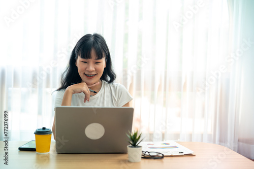 Asian woman sitting wotk happily, using a laptop and enjoying a cup of coffee at a desk with bright natural light. Talking through the laptop screen photo