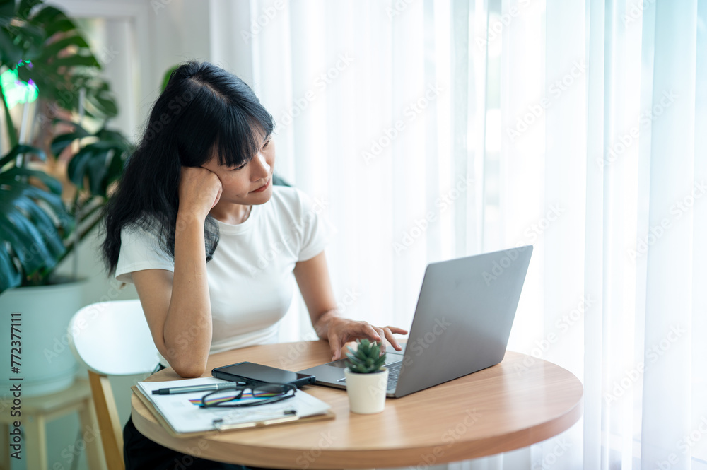 Concentrated Asian woman working by checking data on laptop Serious thinking and planning, smartphone, coffee and documents on the table.