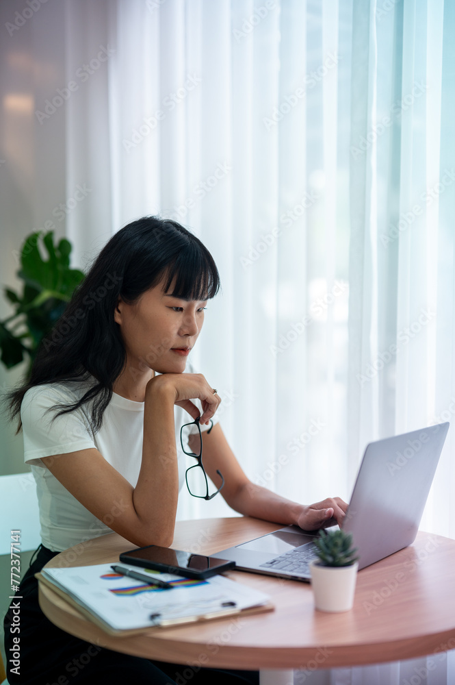 Concentrated Asian woman working by checking data on laptop Serious thinking and planning, smartphone, coffee and documents on the table. Concentrated Asian woman working by checking data on laptop Serious thinking and planning, smartphone, coffee and documents on the table.