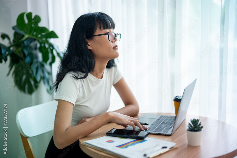 Asian woman wearing reflective glasses looks into the distance thoughtfully, gets bored, and stops working on her laptop in a well-lit room. Asian woman wearing reflective glasses looks into the distance thoughtfully, gets bored, and stops working on her laptop in a well-lit room.