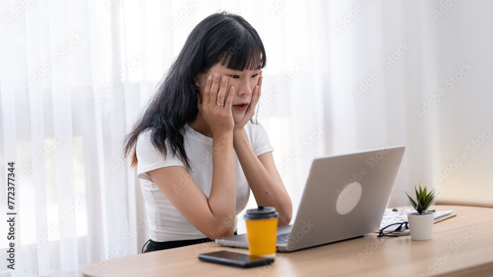 Worried Asian woman looking at laptop screen, expressing stress and concern with smartphone and coffee on desk. Worried Asian woman looking at laptop screen, expressing stress and concern with smartphone and coffee on desk.