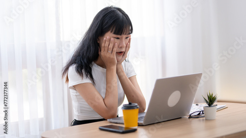 Worried Asian woman looking at laptop screen, expressing stress and concern with smartphone and coffee on desk.