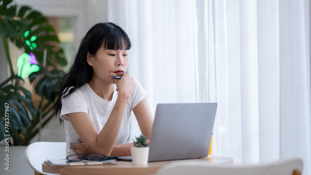 Concentrated Asian woman working by checking data on laptop Serious thinking and planning, smartphone, coffee and documents on the table. Concentrated Asian woman working by checking data on laptop Serious thinking and planning, smartphone, coffee and documents on the table.