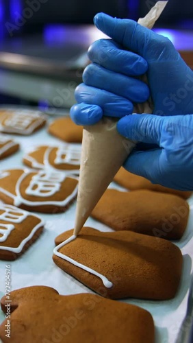 Confectionery worker holding a pastry bag and squeezing white cream on the cookies. Gingerbread houses close up. Trays with pastry in blur at background. Vertical video photo