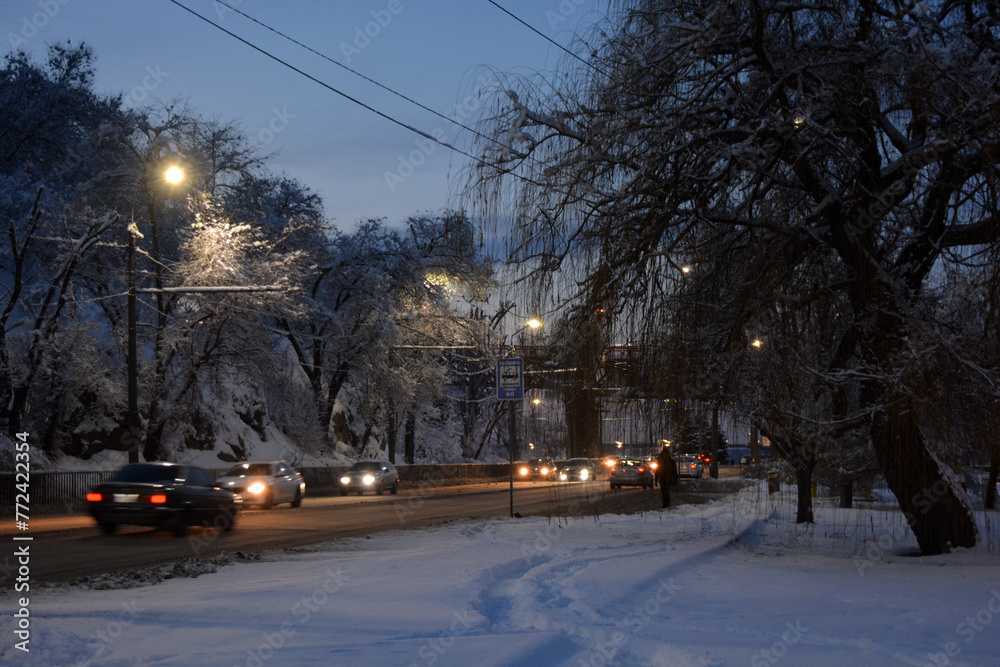 A winter and night city with people, benches, houses, buildings, white snow, bare trees, lanterns are located along the Dnipro River. Very unusual sections of the winter embankment of  Dnipro River.