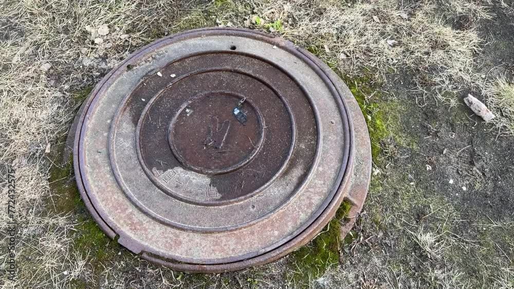 A dirty rusty iron hatch with a pattern is located next to dry grass during the daytime. The water is on the surface of the hatch. Close-up A dirty rusty iron hatch with a pattern is located next to dry grass during the daytime. The water is on the surface of the hatch. Close-up