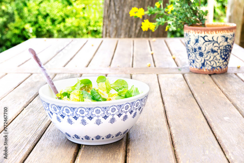 Bowl of fresh salad next to a pot of flowers on a wooden table on the patio.