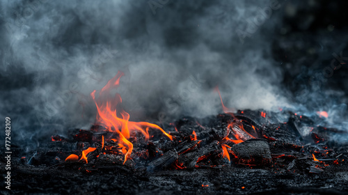 The dying flames of a fire are smoldering amongst charcoal and ash, with wisps of smoke curling up into the cool air.