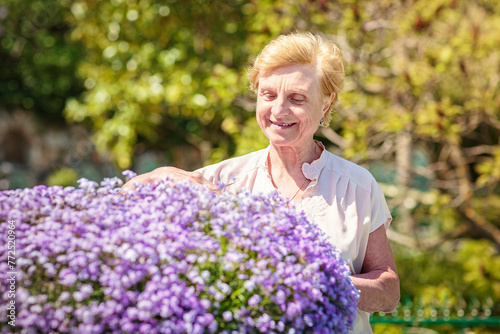 Elderly woman standing near a large flower pot with purple flowers and caring for plants