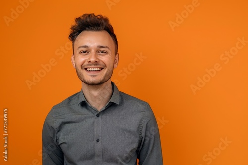 A man in a gray shirt stands before an orange wall, grinning broadly