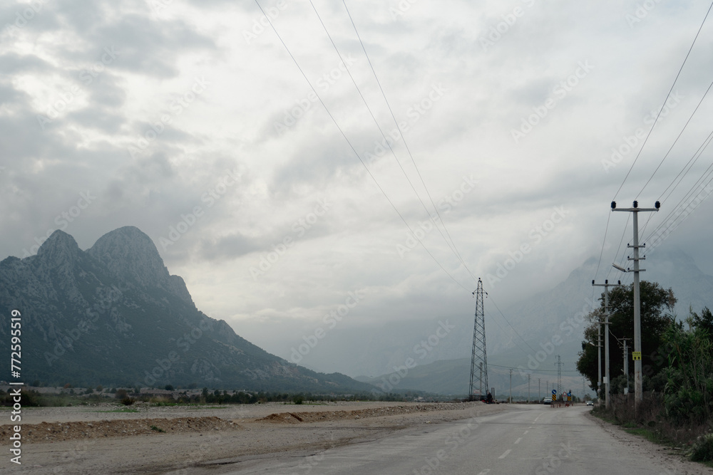 A two-lane asphalt road stretches towards a mountain in the distance. Power lines run along the side of the road