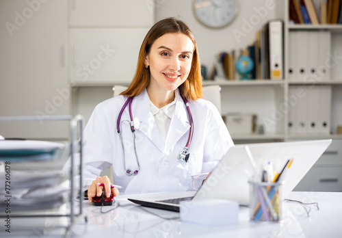 Young woman doctor sitting at table in her office and looking at camera.
