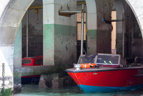 A vivid red boat moored in the shadows of an underpass, with the contrasting weathered concrete pillars offering a glimpse into Venice's utilitarian spaces