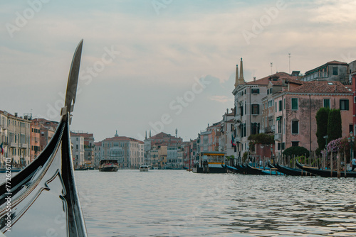 The prow of a gondola cuts through the serene waters of Venice's Grand Canal at dusk, with historic pastel buildings and boats lining the sides.