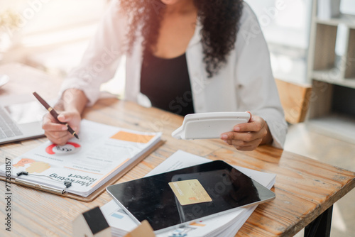 Focused businesswoman using a calculator and reviewing financial reports, surrounded by various work documents on her desk.