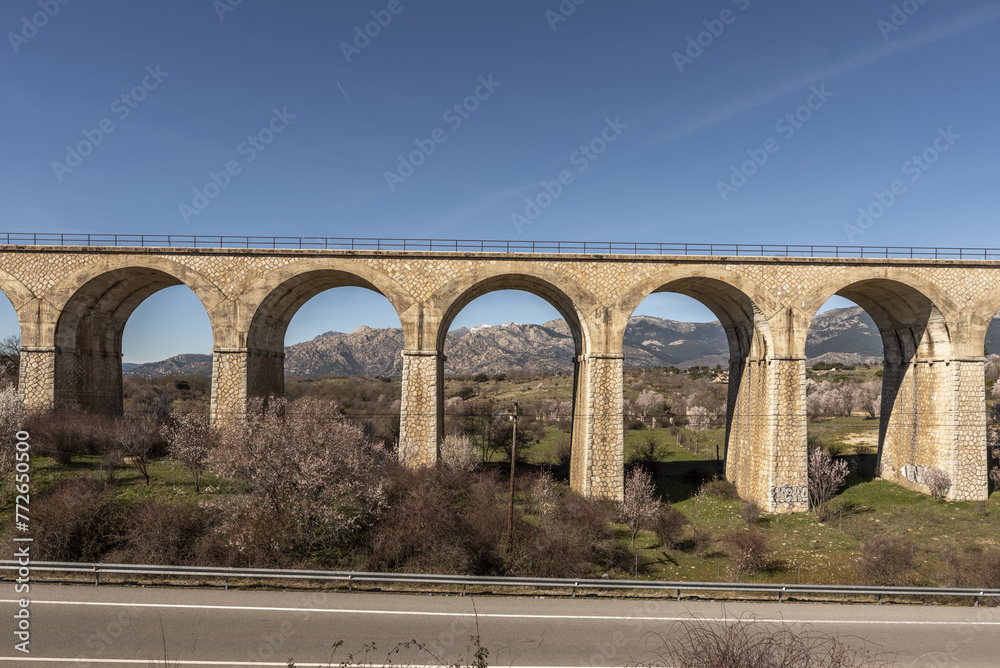 An old stone railway bridge with many arches through which you can see a mountain range next to a conventional road.