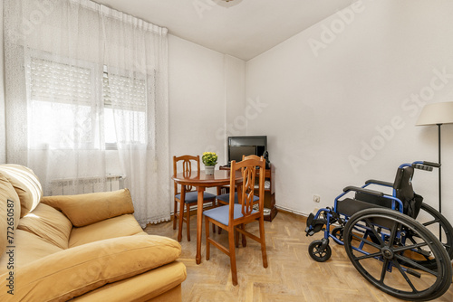 Corner of a small living room with round wooden table with matching chairs upholstered in blue microfiber with a black wheelchair next to it