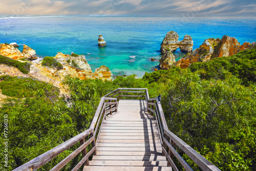 Scenic Wooden Boardwalk Leading to Turquoise Sea, Lush Greenery, and Rugged Cliffs at Coastal Destination
