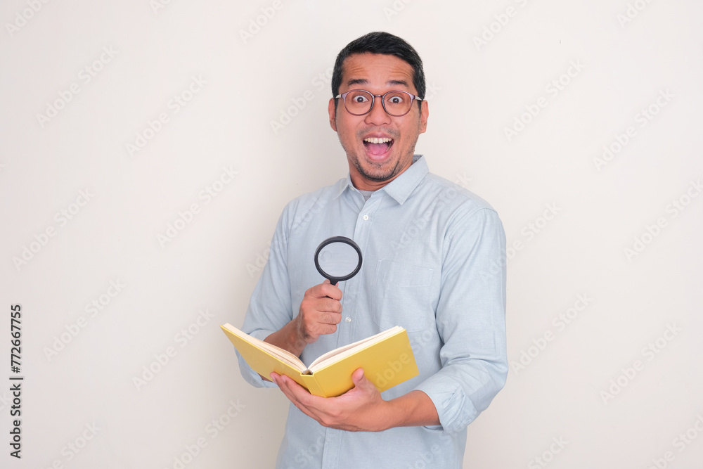 Adult Asian man showing wow face expression while holding a book and magnifying glass Adult Asian man showing wow face expression while holding a book and magnifying glass