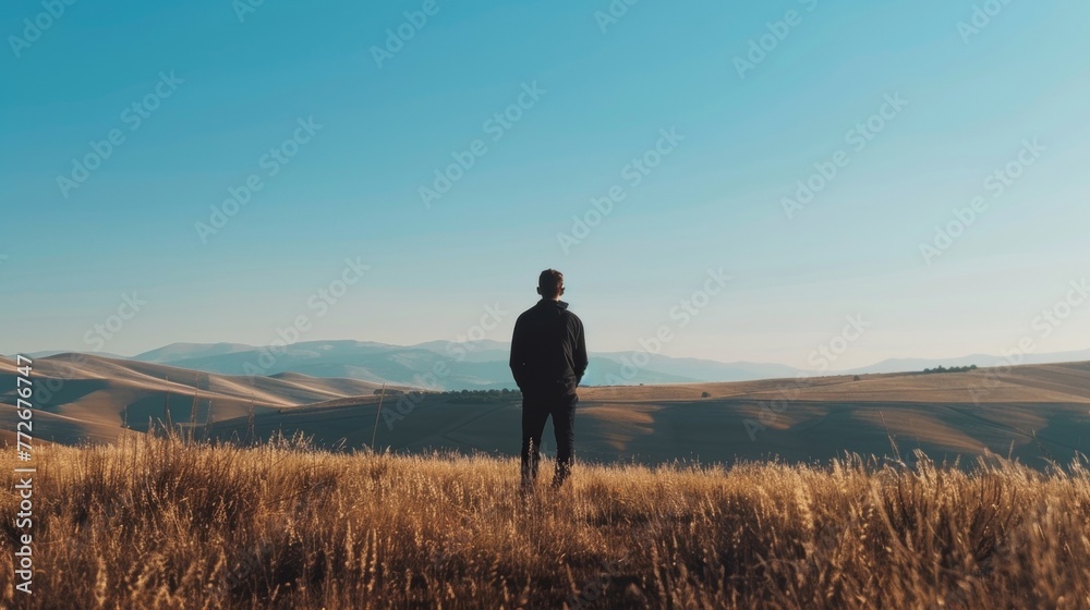 Against a backdrop of rolling hills and clear skies a figure stands with back to the camera enjoying a moment of solitude and . . Against a backdrop of rolling hills and clear skies a figure stands with back to the camera enjoying a moment of solitude and . .