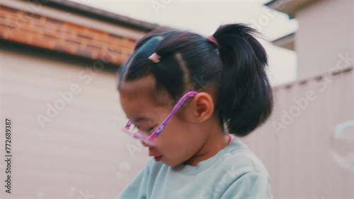 Little pretty asian girl wearing glasses is playing outdoor in front of garage with happy face in summer and happily running around with soap bubbles.	4k Resolution.