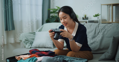 Selective focus of Asian teenager woman wear wireless headphones sitting on sofa look pictures on the camera while packing suitcase luggage in the living room , Preparation travel suitcase at home.