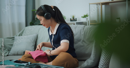 Selective focus of Asian teenager woman wear wireless headphones sitting on sofa packing travel luggage with clothes for traveling trip, Preparation travel suitcase at home.