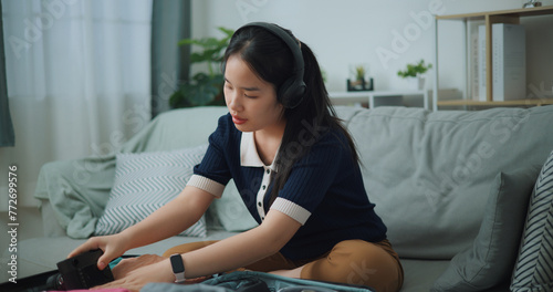 Selective focus of Asian teenager woman wear wireless headphones sitting on sofa packing travel luggage with personal items for traveling trip, Preparation travel suitcase at home.
