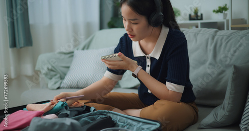Selective focus of Asian teenager woman wear wireless headphones sitting on sofa making checklist of things to pack for travel, Preparation travel suitcase at home.