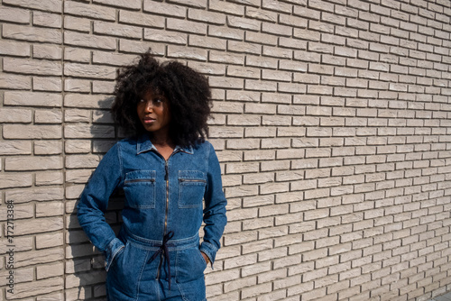 A stylish young African American woman with a voluminous afro hairstyle stands casually against a textured brick wall, her hands tucked into the pockets of her chic denim jumpsuit. Her sideways glance photo