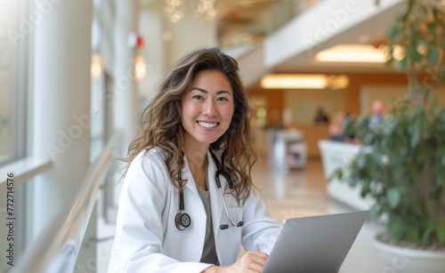 Friendly female doctor working on a laptop in a well-lit hospital lobby, showcasing modern healthcare practices