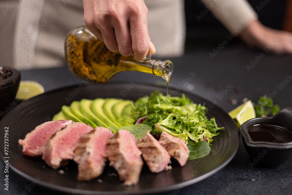Woman pours cooking olive oil on a salad on a plate with grilled cooked piece of tuna fillet