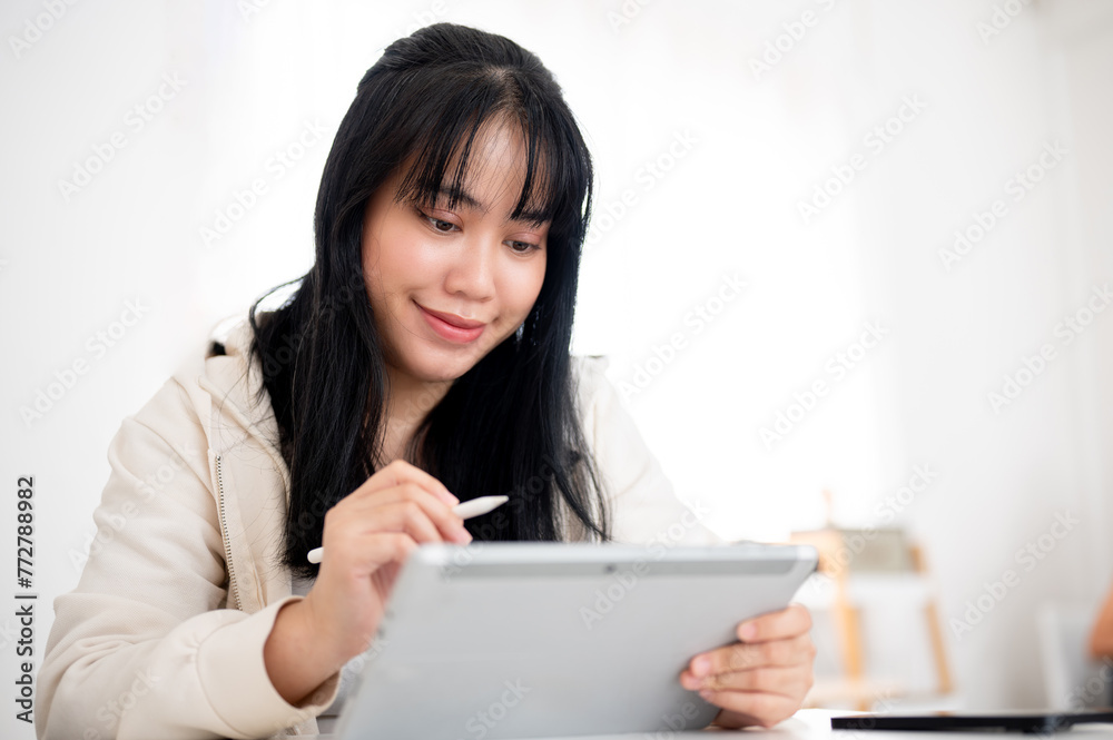 A young Asian woman using her digital tablet with a stylus pen at a table indoors. A young Asian woman using her digital tablet with a stylus pen at a table indoors.