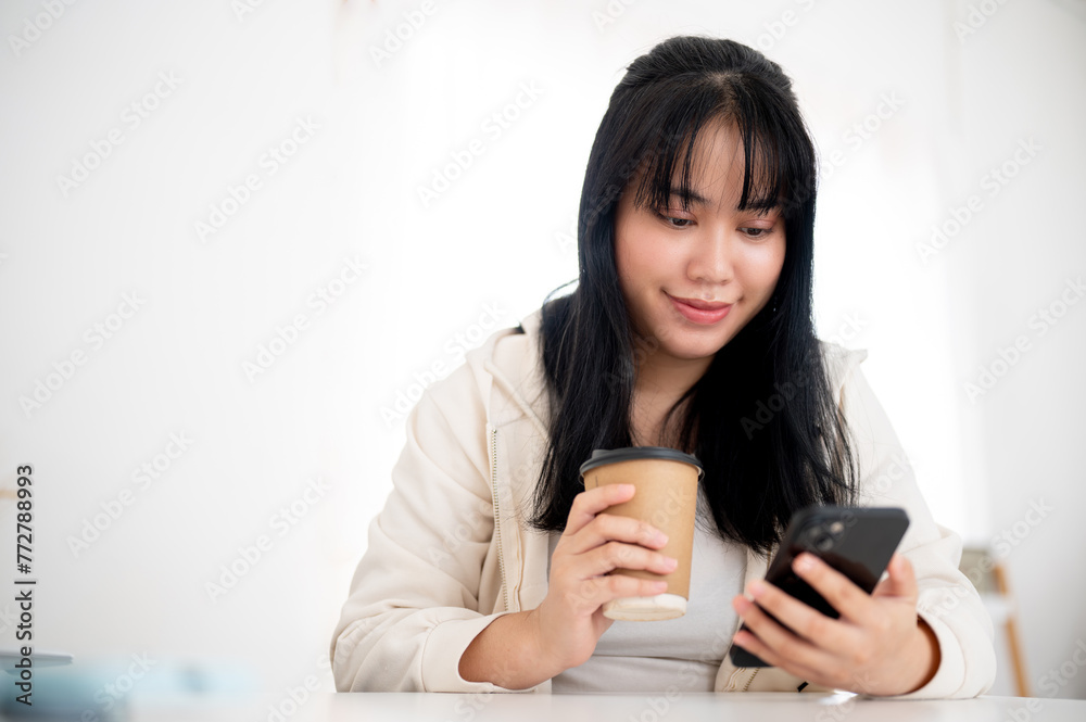 A positive, smiling Asian woman in casual wear is sipping coffee and using her smartphone in a cafe. A positive, smiling Asian woman in casual wear is sipping coffee and using her smartphone in a cafe.