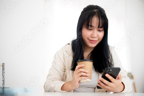 A positive, smiling Asian woman in casual wear is sipping coffee and using her smartphone in a cafe.