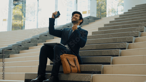 Successful business man celebrate successful project while sitting at stairs. Smart project manager getting new gob, getting promotion, increasing sales while calling friends by using phone. Exultant. photo