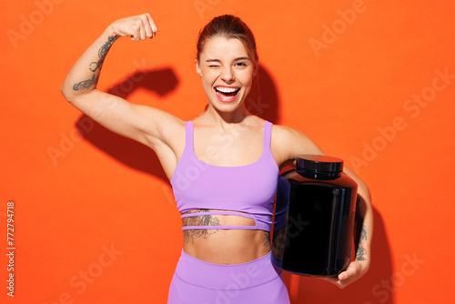 Young cool fitness trainer instructor woman sportsman wear purple top clothes in home gym hold bottle of supplements wink show biceps isolated on plain orange background Workout sport fit abs concept.