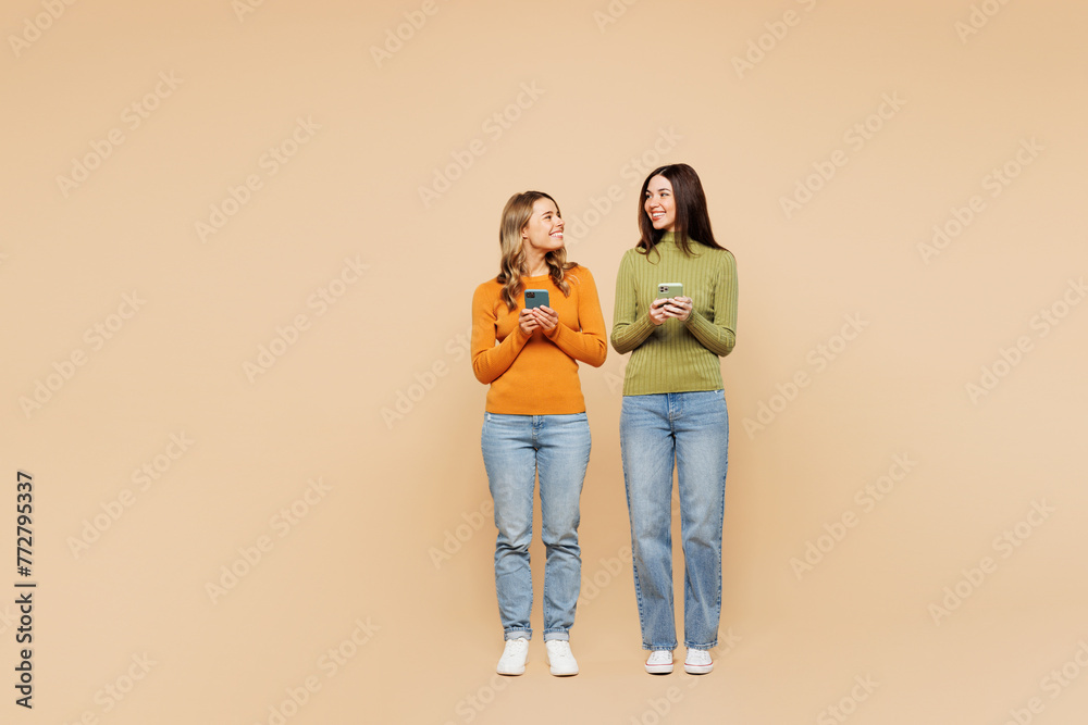 Full body young friends two women wearing orange green shirt casual clothes together hold in hand use mobile cell phone look to each other isolated on plain pastel beige background. Lifestyle concept. Full body young friends two women wearing orange green shirt casual clothes together hold in hand use mobile cell phone look to each other isolated on plain pastel beige background. Lifestyle concept.