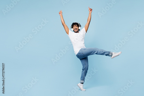 Full body young happy Indian man wear white t-shirt casual clothes listen to music in headphones raise up hands isolated on plain pastel light blue cyan background studio portrait. Lifestyle concept. photo