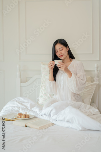 A beautiful Asian woman in white long dress pajamas is having breakfast on bed, sipping tea.