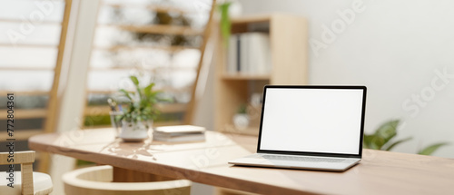 A white-screen laptop mockup on a wooden dining table in a neutral contemporary dining room. © bongkarn