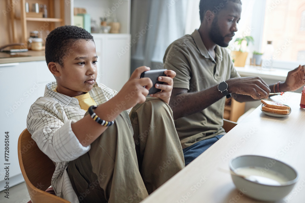Portrait of teenage African American boy using smartphone at kitchen table during breakfast with family copy space Portrait of teenage African American boy using smartphone at kitchen table during breakfast with family copy space