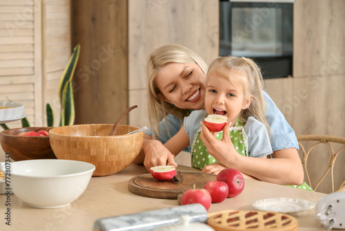 Little girl with her mother eating cut apple while cooking pie in kitchen