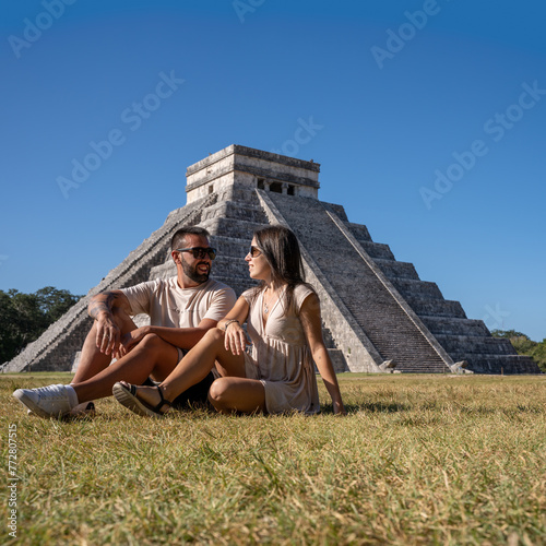 Lovely couple in love visiting Chichen Itza, sitting on the grass with the pyramid in the background.