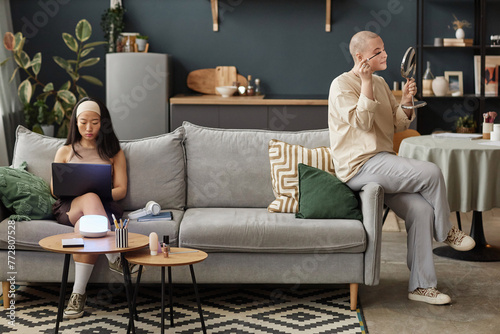 Wide shot of two diverse women spending time at home sitting on sofa in living room surfing internet on laptop and doing makeup, copy space
