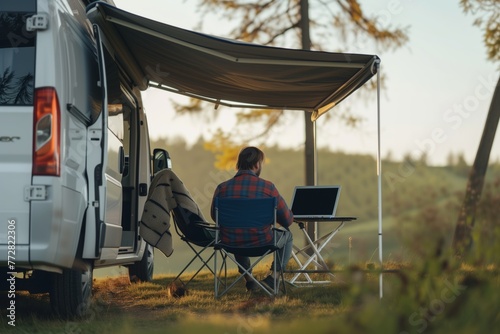 person sitting under awning attached to camper van with a laptop