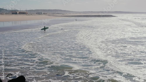 Aerial view of man carrying surfing board and go into ocean for surfing at sunny summer day. Male surfer in sea water with waves. Water sport and hobby concept. Active lifestyle