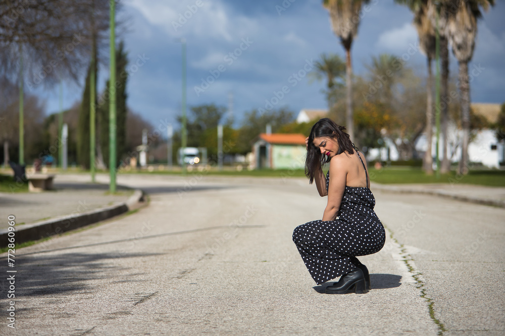 A young, beautiful woman in a black dress with white polka dots, crouched down, sad and alone, in the middle of a road. Concept loneliness, sorrow, beauty, fashion.